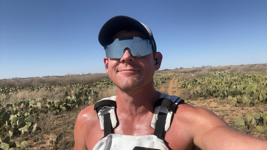 Preston Shamblen rucking solo through a cactus-lined dirt trail in West Texas, wearing a white Wolf Tactical vest and reflective sunglasses under a clear blue sky.