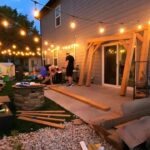 Two men assembling the cedar frame of a Backyard Discovery Arcadia gazebo on a patio at dusk with string lights overhead