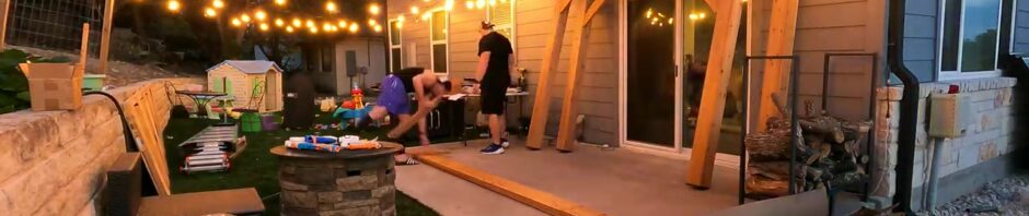 Two men assembling the cedar frame of a Backyard Discovery Arcadia gazebo on a patio at dusk with string lights overhead